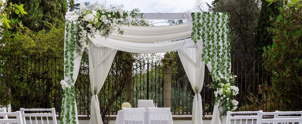 Wedding arch decorated with white flowers and artificial ivy garlands, with white chairs in the foreground.