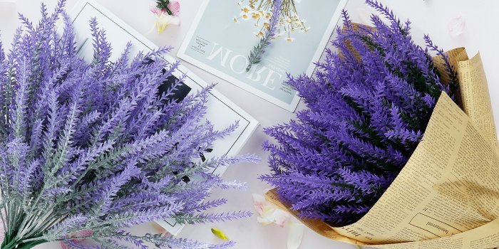 Two bundles of artificial lavender flowers, one wrapped in newspaper, on a light pink surface with magazines and decorative elements.