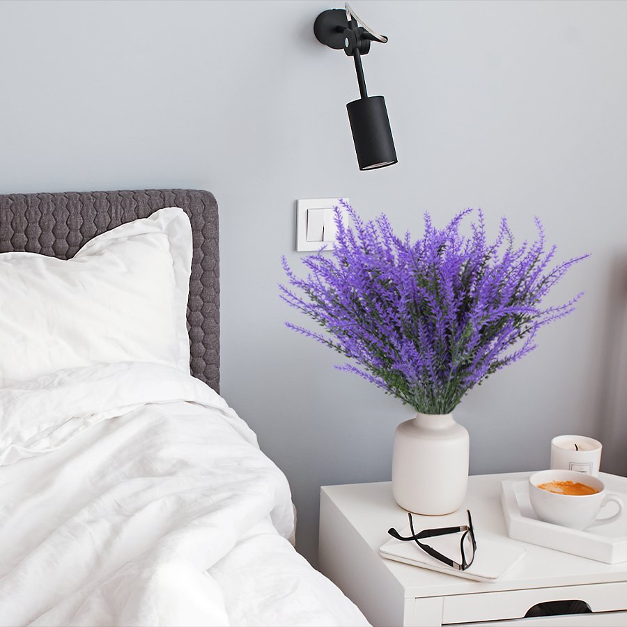 Artificial lavender bundles in a vase on a nightstand next to a bed.