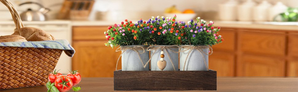 Artificial flower centerpiece with three metal pots on a wooden tray, set on a kitchen counter with a basket of bread and tomatoes.