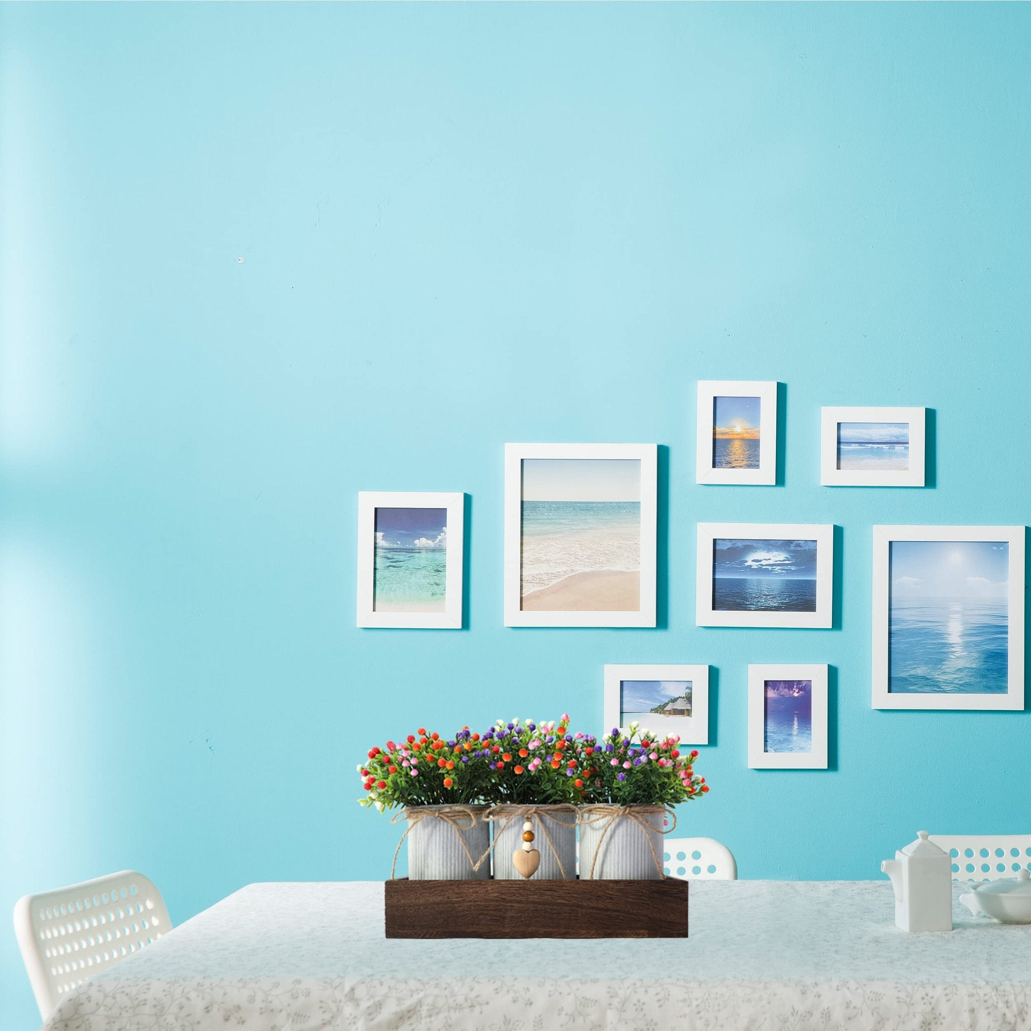 Artificial flower centerpiece with three metal pots in a wooden tray, set on a tablecloth with framed beach photos on a blue wall.