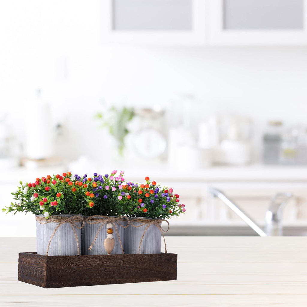 Artificial flower centerpiece with three metal pots in a wooden tray, set on a white wooden surface with a blurred kitchen background.