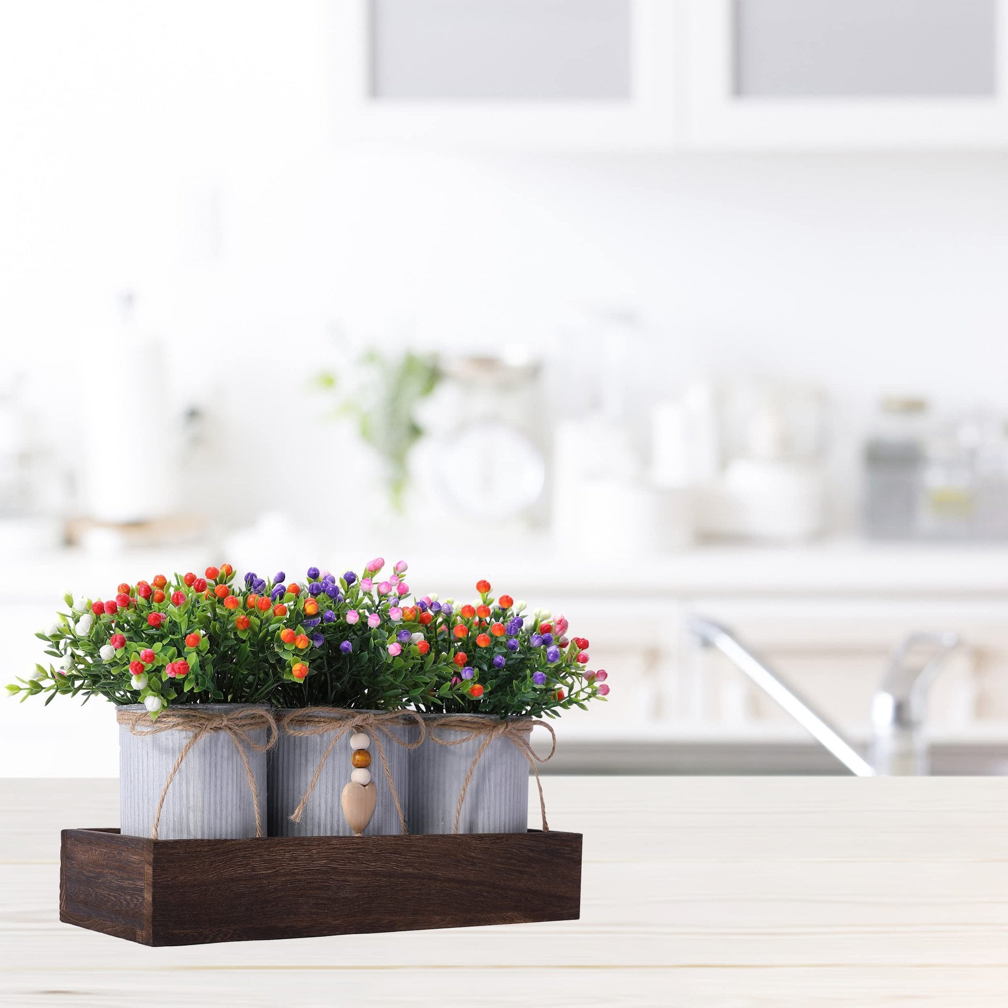Artificial flower centerpiece with three metal pots in a wooden tray, set on a white wooden surface with a blurred kitchen background.