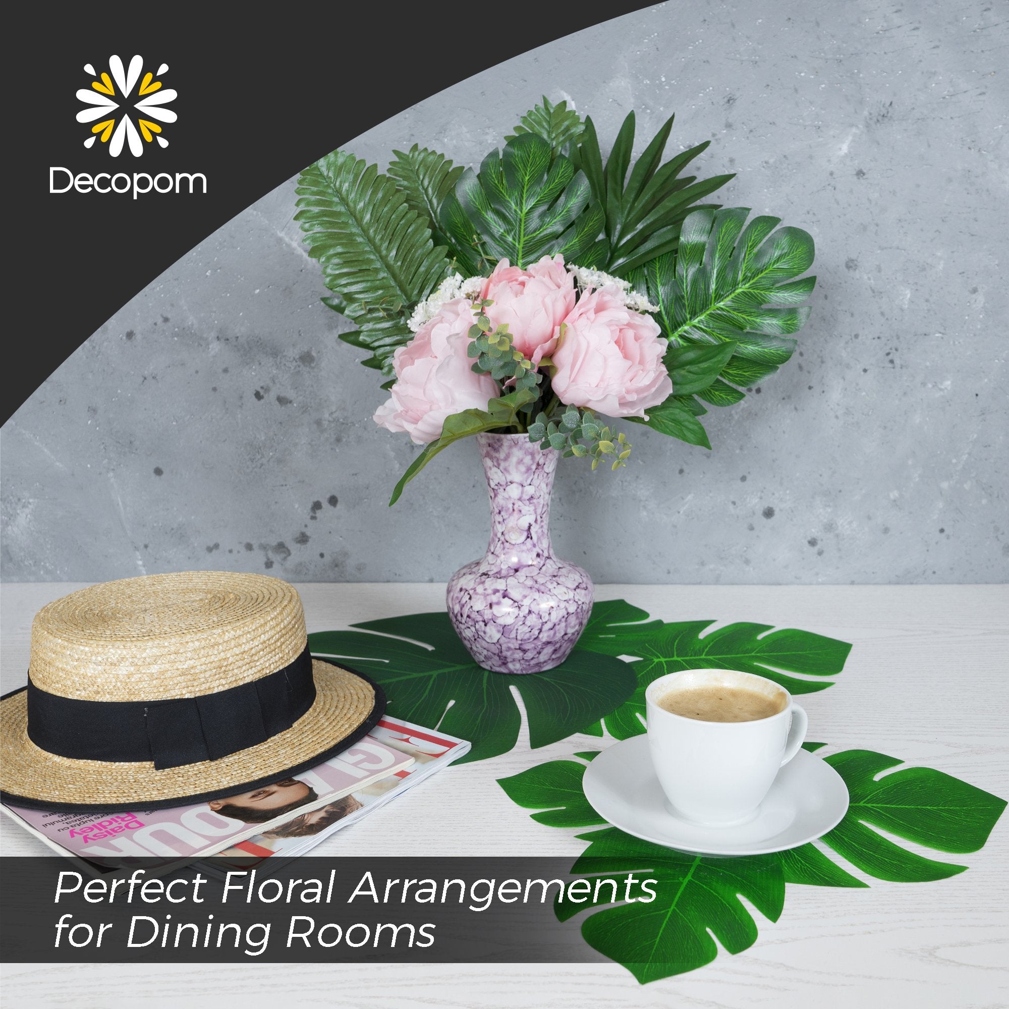 Artificial palm leaves arranged with a vase of pink flowers, a straw hat, and a cup of coffee on a table.