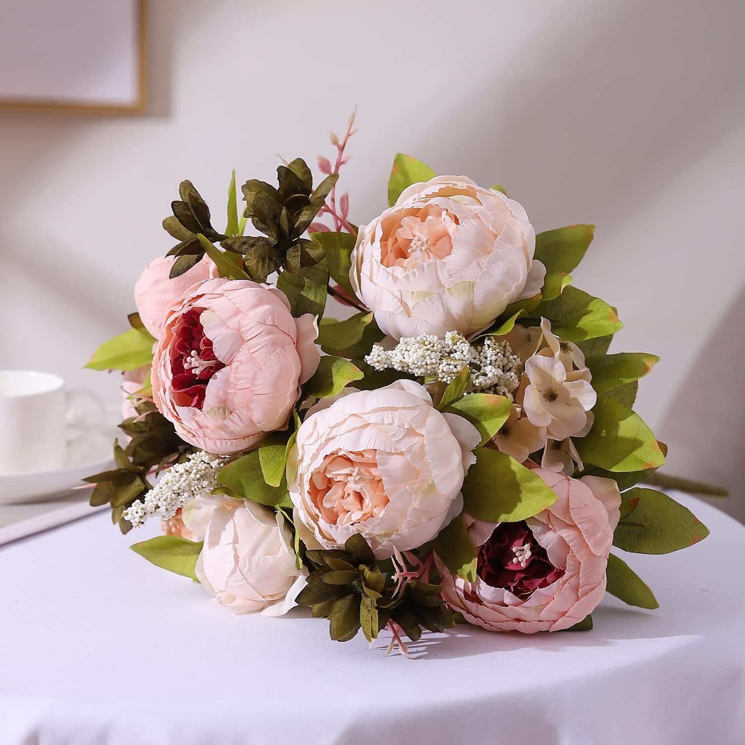 Light pink artificial peony flower bouquet with buds and green leaves on a white tablecloth.