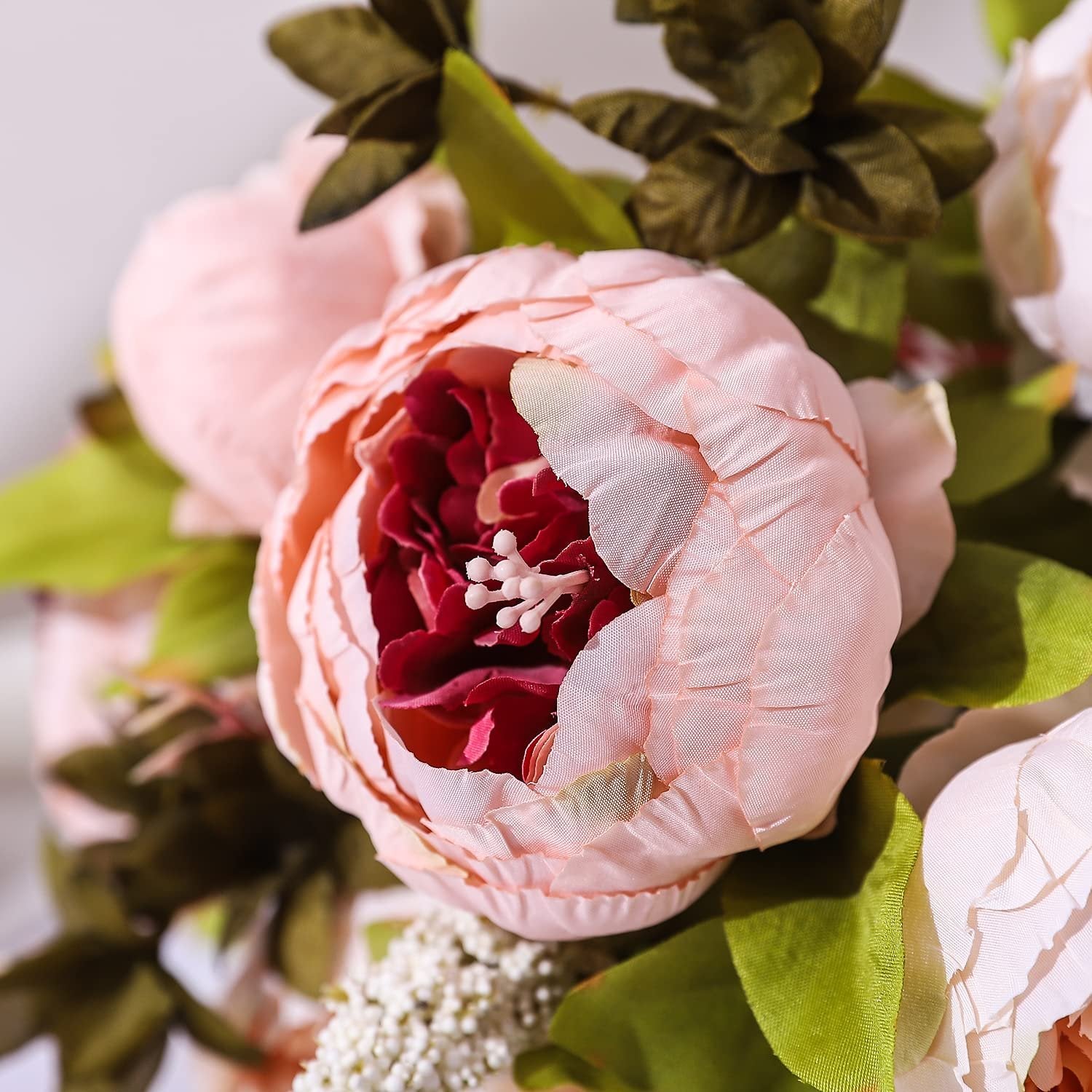 Close-up of a light pink artificial peony flower with a dark red center and green leaves.