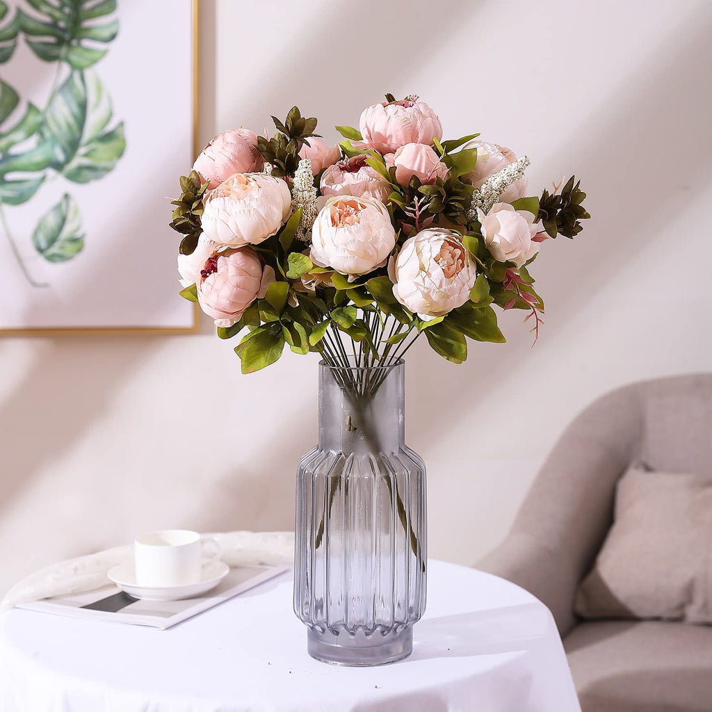 Light pink artificial peony flower bouquet in a ribbed glass vase on a white tablecloth, with a framed plant artwork in the background.