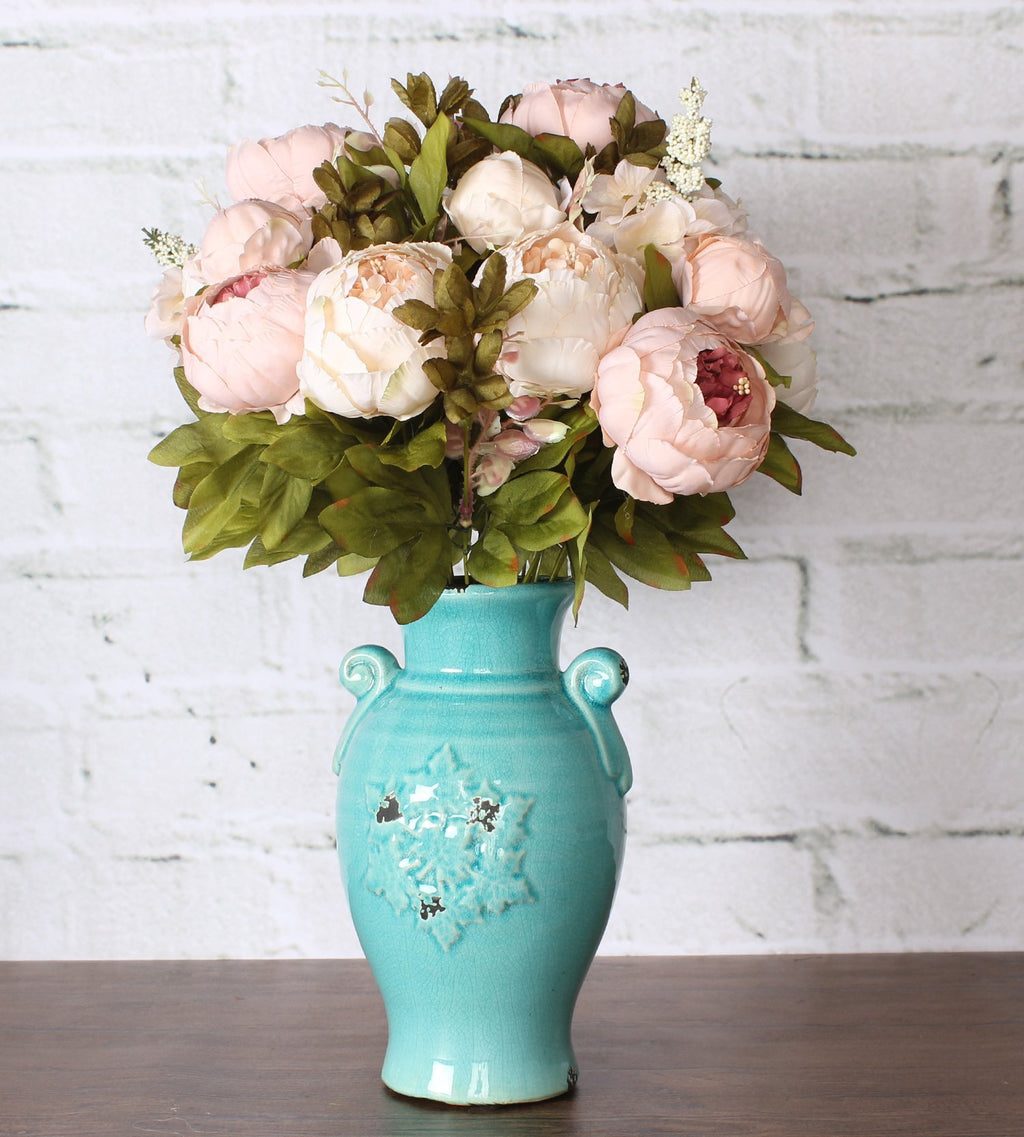Light pink peony silk flower bouquet with green leaves in a turquoise vase against a white brick background.