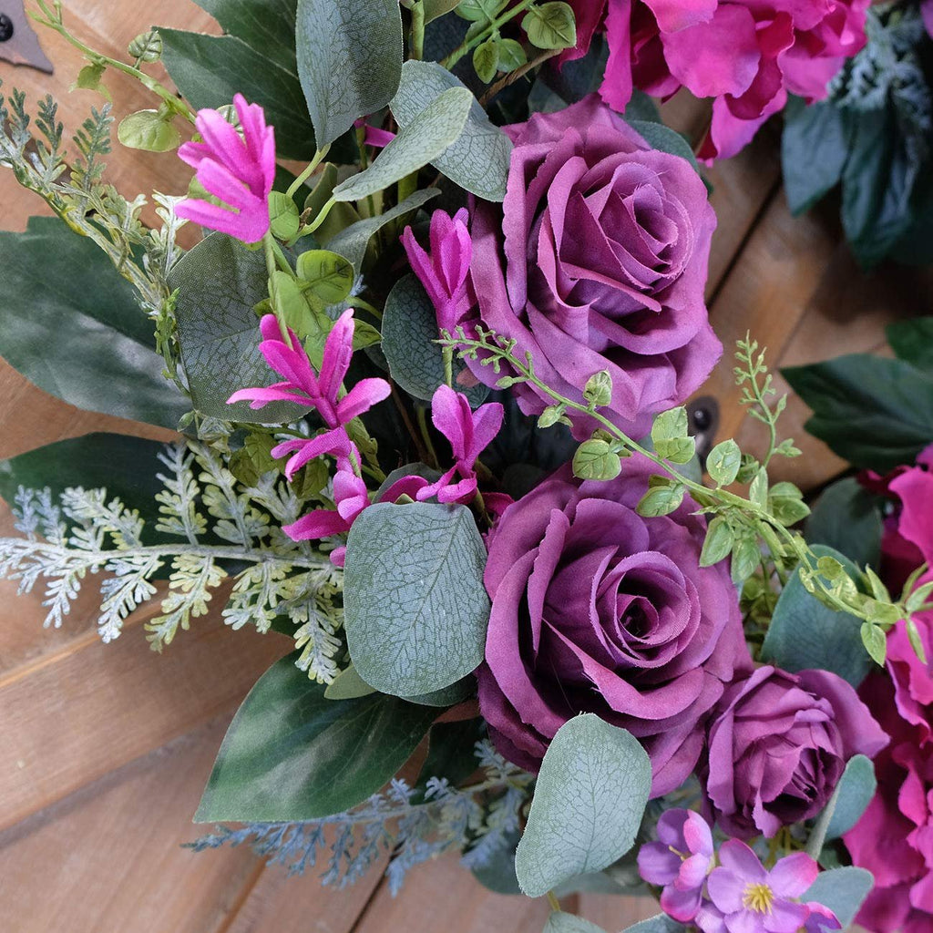 Close-up of an artificial purple rose wreath with silk hydrangea and eucalyptus leaves on a wooden surface.