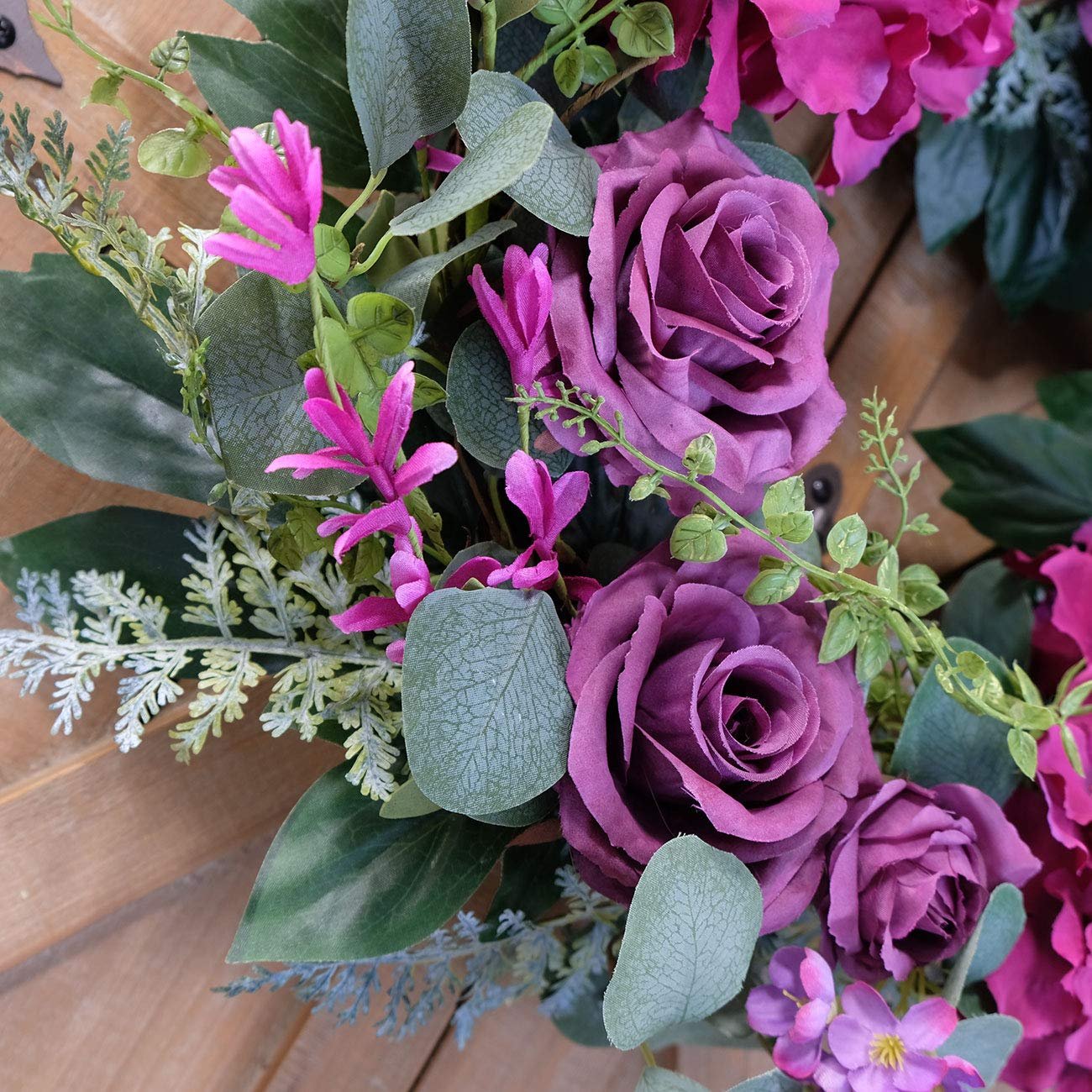 Close-up of an artificial purple rose wreath with silk hydrangea and eucalyptus leaves on a wooden surface.