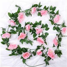 Artificial pink rose vine garland with green leaves draped on a white cloth background, showing multiple flowers and foliage.