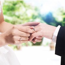 Bride places a ring on groom's finger during a wedding ceremony with blurred outdoor greenery in the background.
