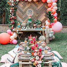 Outdoor party setup with low tables, cushions, balloons, and a dessert table featuring a cake.