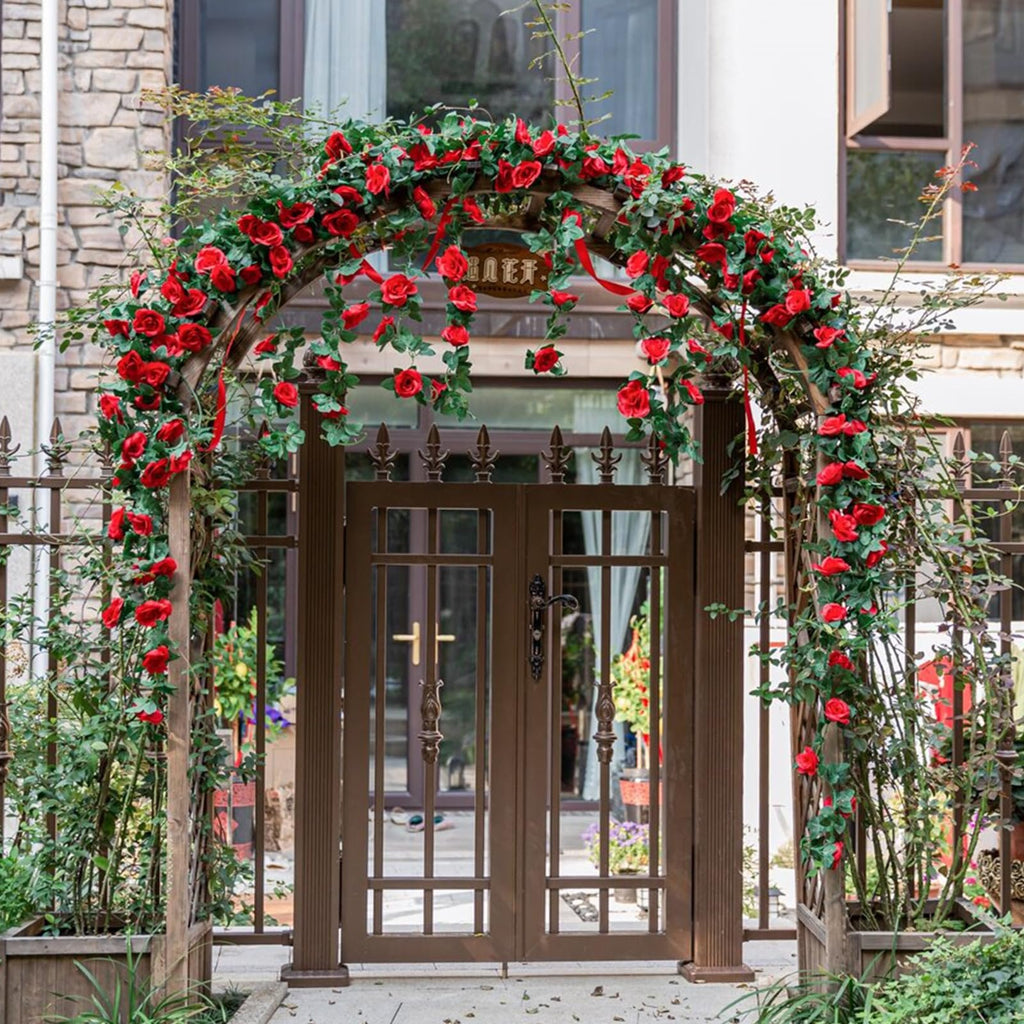 Artificial red rose vine garland draped over a wooden archway in front of a gate, with red ribbons woven through the roses.