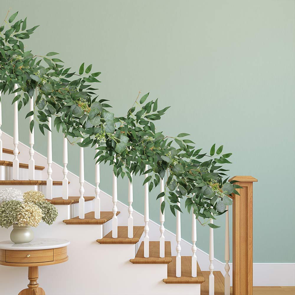 Artificial eucalyptus and willow twig garland draped over a white staircase railing with a vase of flowers on a side table.