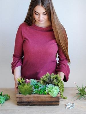 Woman holding a wooden crate filled with assorted artificial succulent plants, with more succulents and decorative stones on a wooden surface.