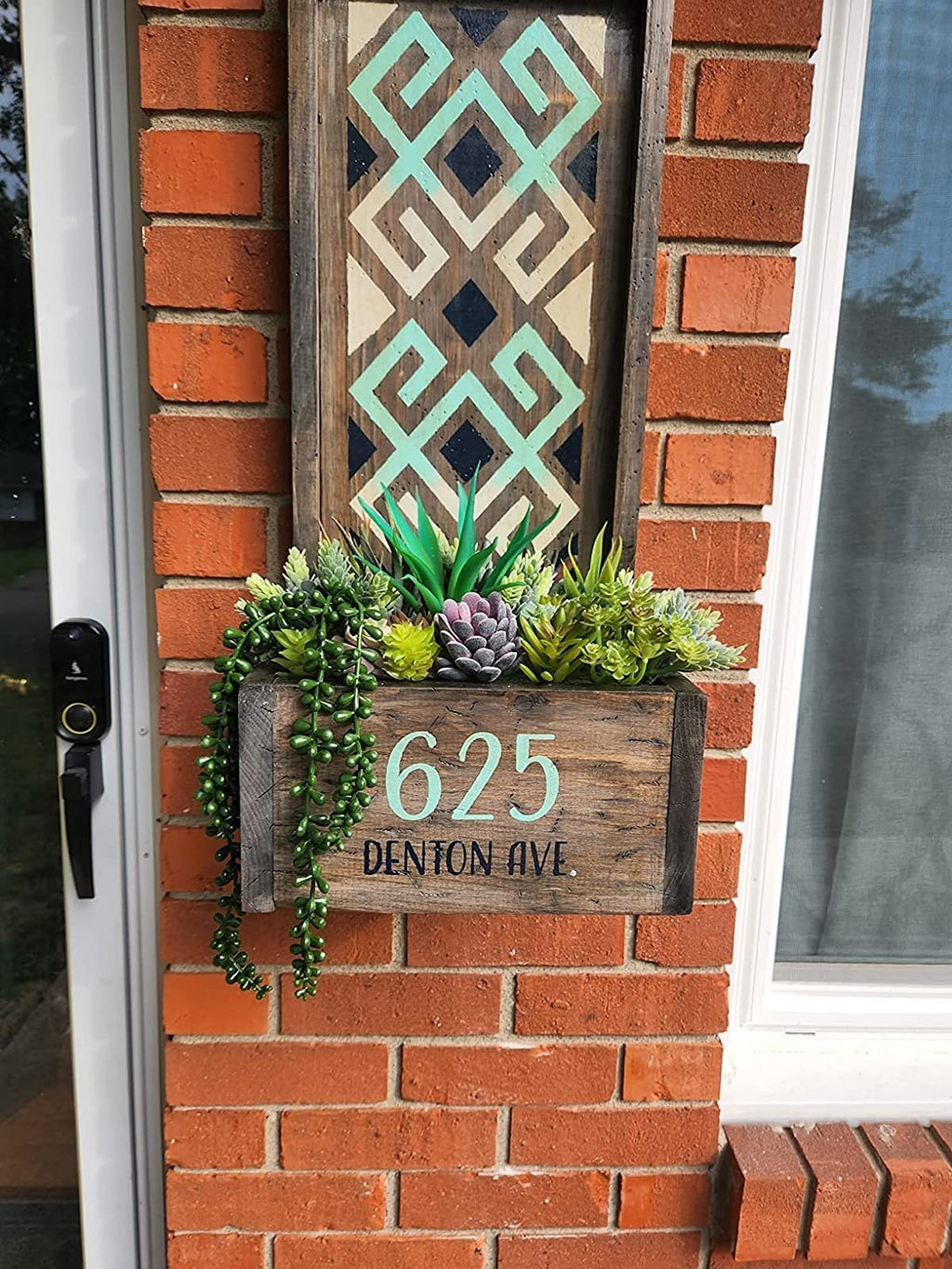 Decorative planter box with artificial succulents mounted on a brick wall next to a door and window, displaying house number 625 DENTON AVE.