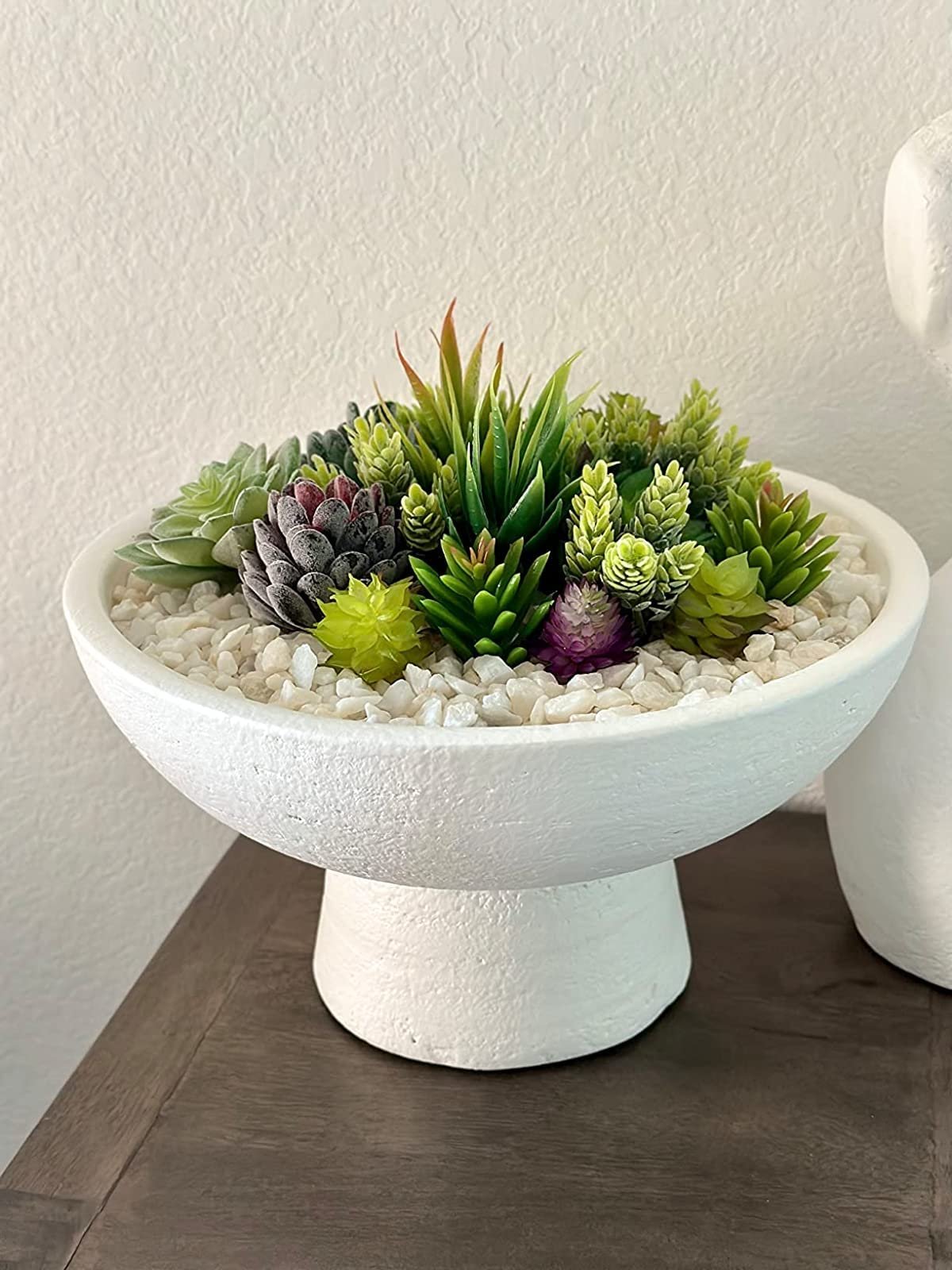 Assortment of unpotted artificial succulents in a white pedestal bowl filled with white pebbles.