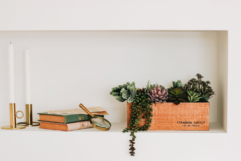 Assorted artificial succulents in a wooden planter box on a shelf with books and candles