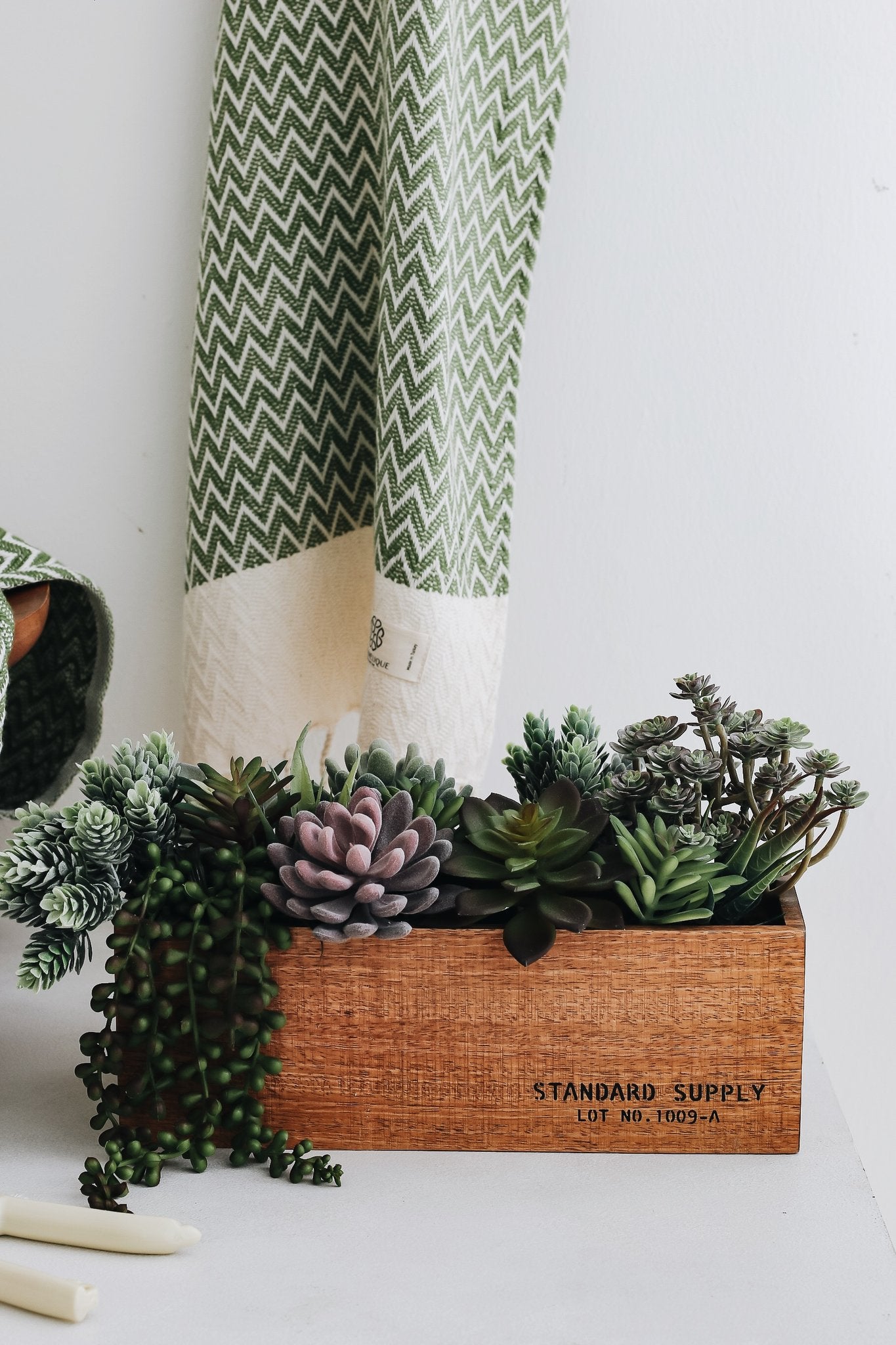 Assorted artificial succulents in a wooden planter box with a green and white patterned towel in the background.