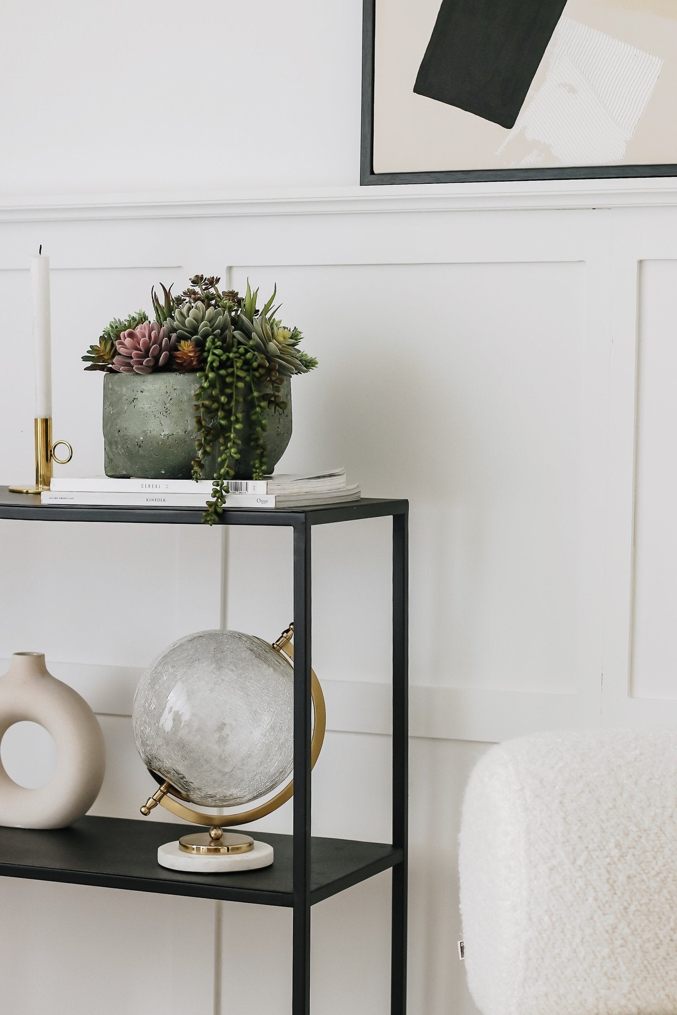 Assorted artificial succulents in a textured gray pot on top of books on a black shelf, next to a candle and a globe.