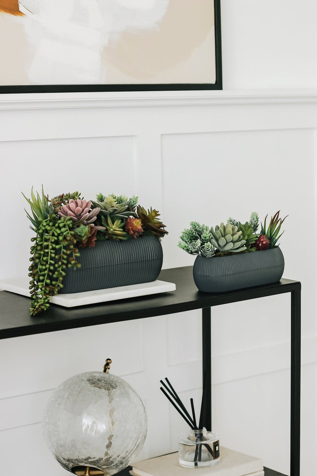 Two decorative planters filled with assorted artificial succulents on a black shelf.