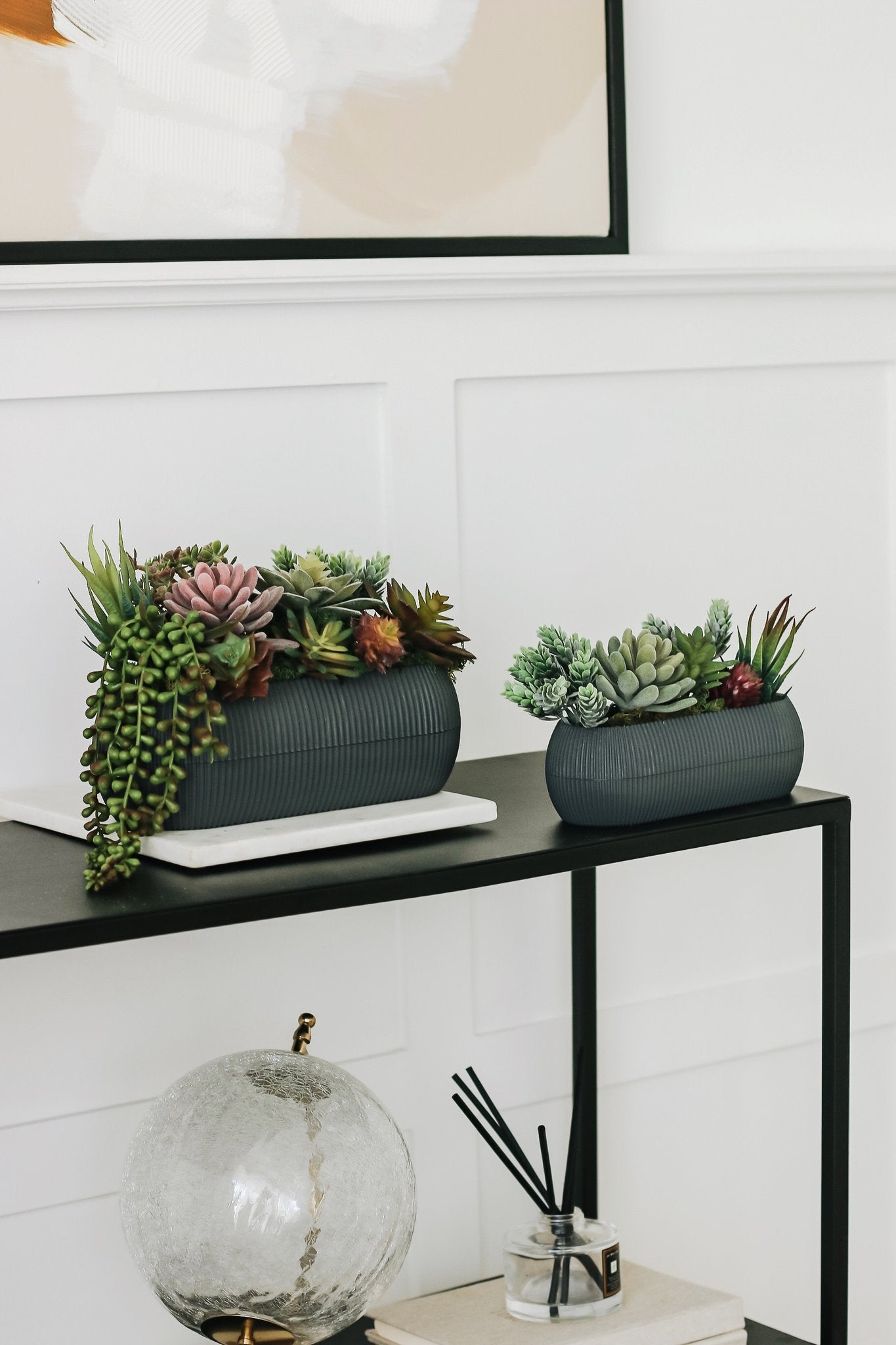 Two decorative planters filled with assorted artificial succulents on a black shelf.