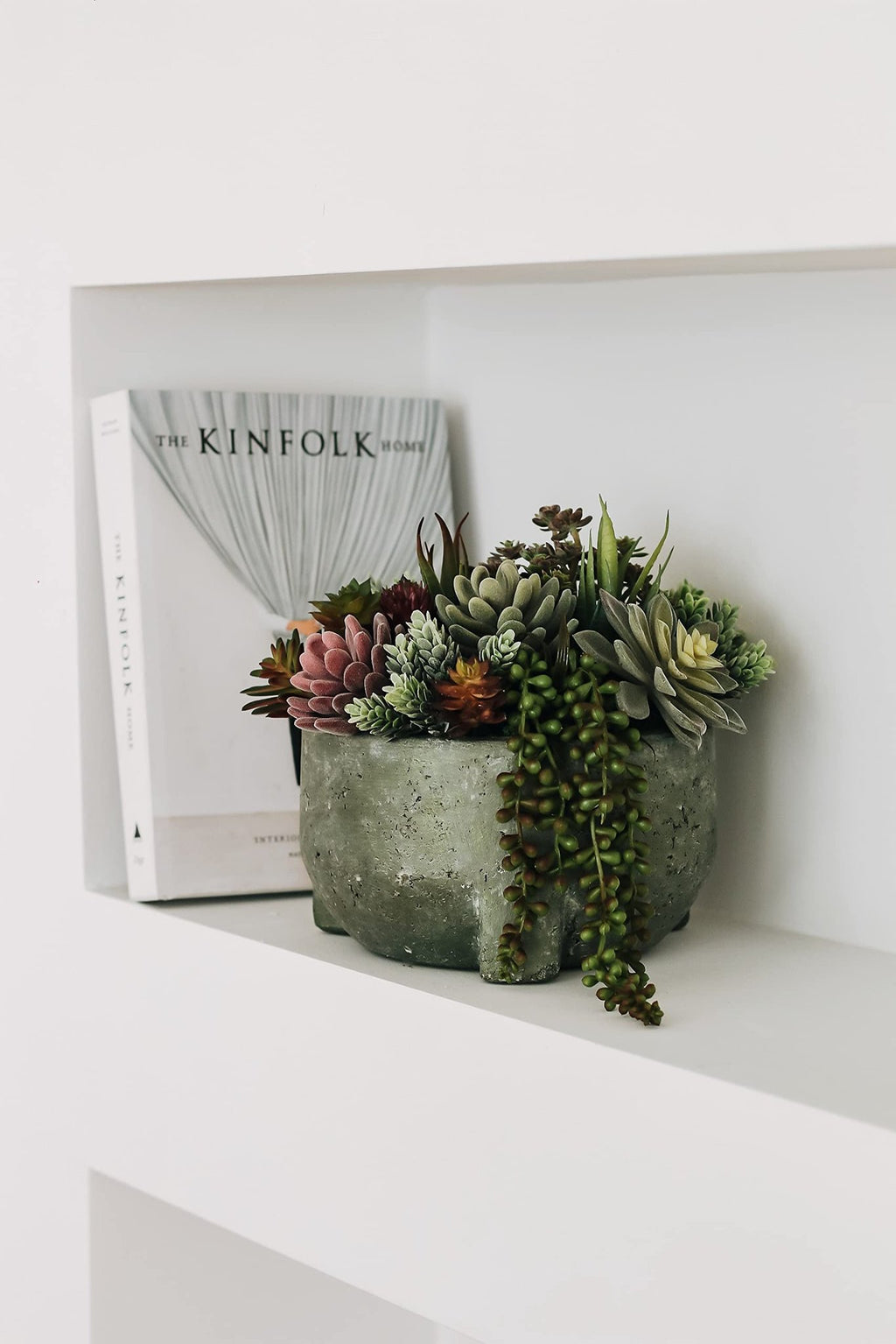 Assorted artificial succulents in a textured gray pot next to a book on a white shelf