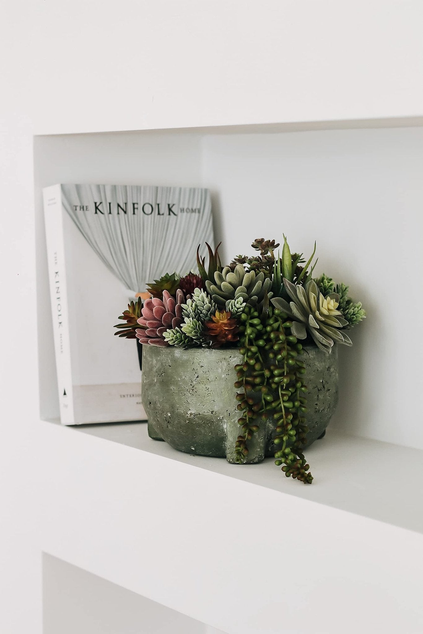 Assorted artificial succulents in a textured gray pot next to a book on a white shelf
