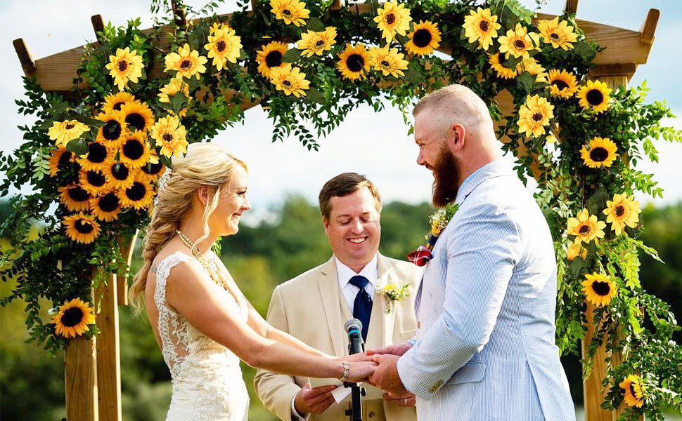 Wedding ceremony with a bride and groom holding hands under a wooden arch decorated with artificial sunflowers and greenery.