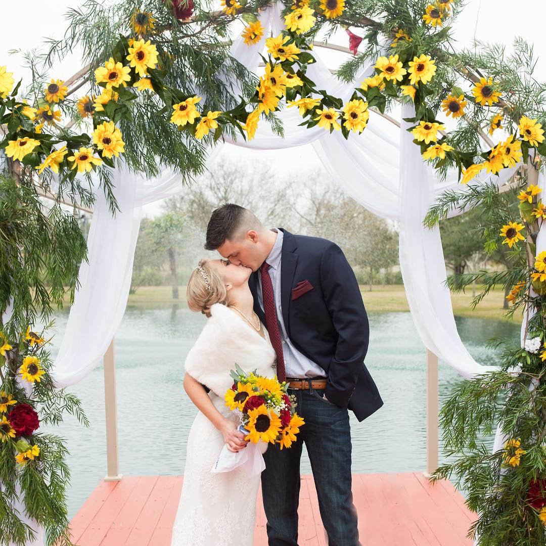 Wedding couple kissing under an arch decorated with artificial sunflowers and greenery, with a lake in the background.
