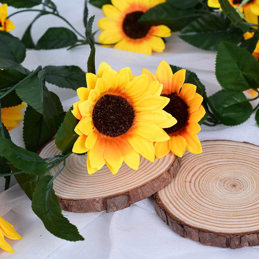 Artificial sunflower garland with green leaves draped over wooden slices on a white cloth background.