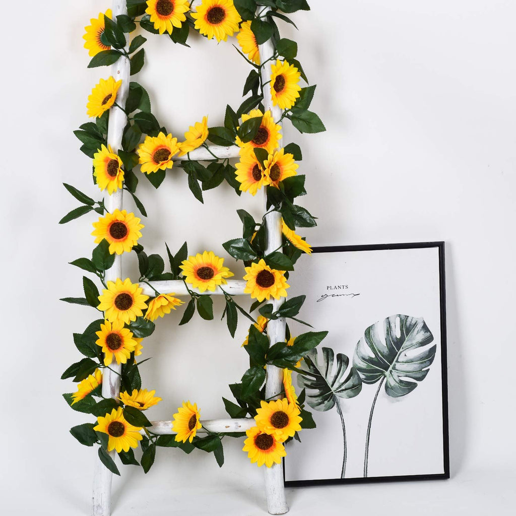 Artificial sunflower garland with green leaves draped on a white ladder next to a framed print of monstera leaves.