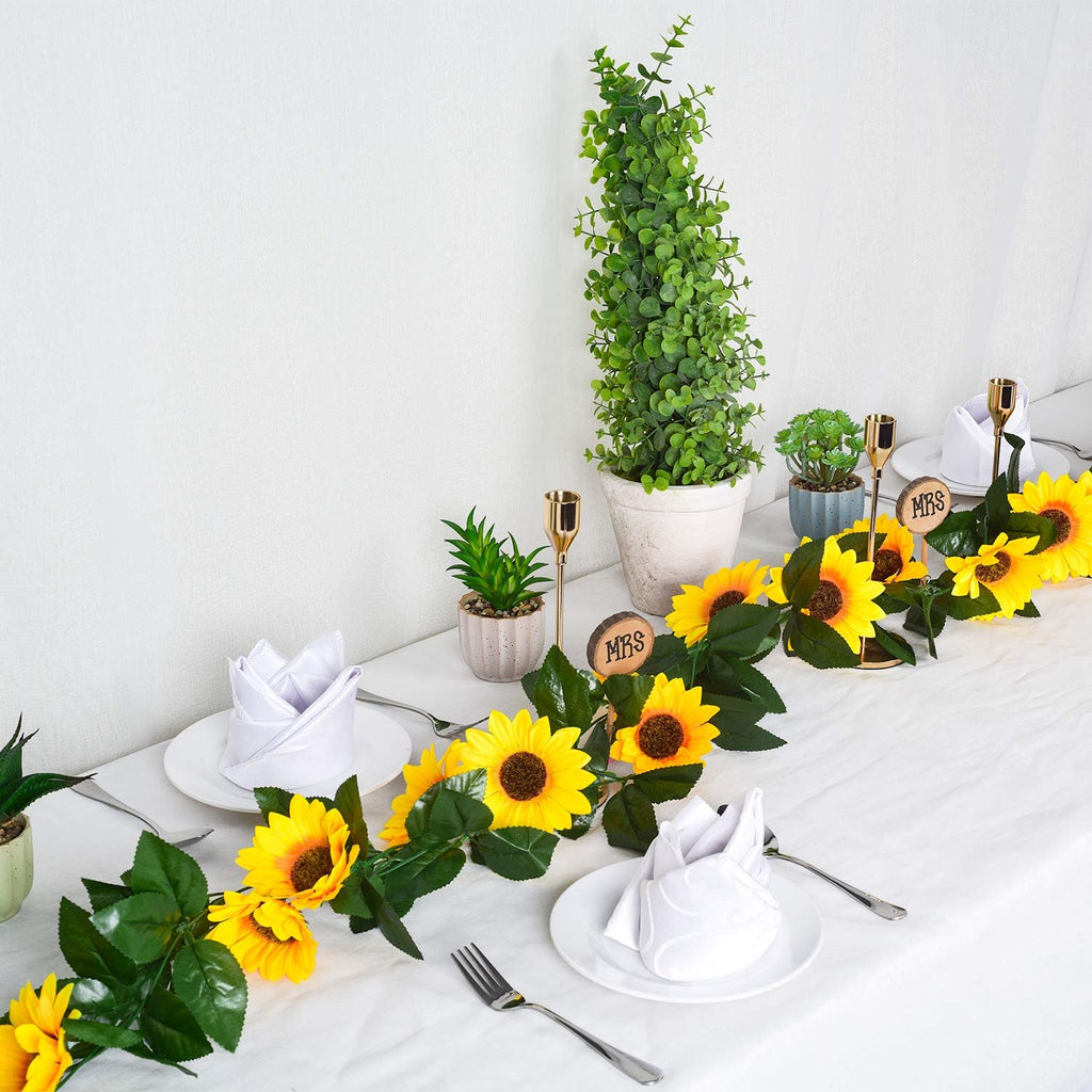 Artificial sunflower garland with green leaves draped across a white tablecloth with place settings and potted plants.