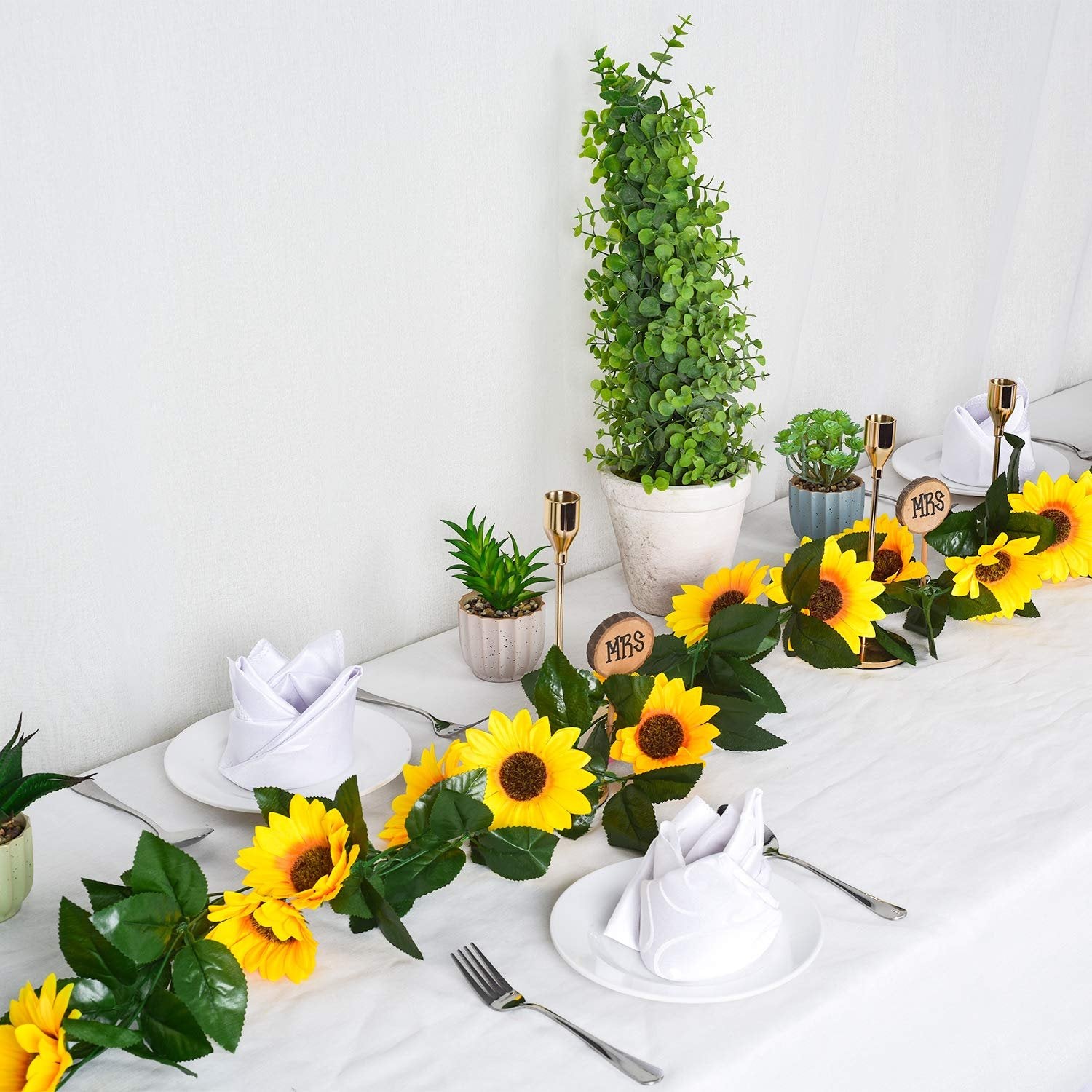 Artificial sunflower garland with green leaves draped across a white tablecloth with place settings and potted plants.