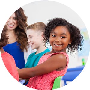 Smiling Black girl in pink dress with teacher and classmates in background during an art class activity.