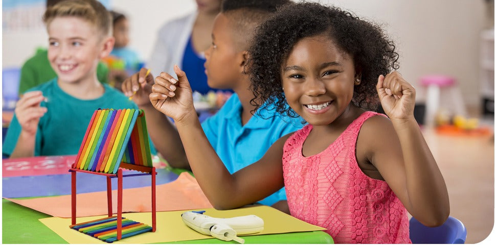 Children engaged in arts and crafts, building a colorful popsicle stick structure. A girl in a pink dress smiles at the camera.