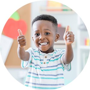 Young boy giving two thumbs up with a big smile, wearing a striped t-shirt, in a classroom setting.