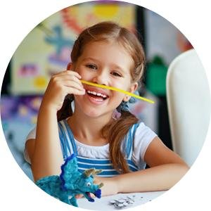 Young girl with pigtails smiling and holding a yellow craft stick to her mouth, with a blue pipe cleaner dinosaur on the table.