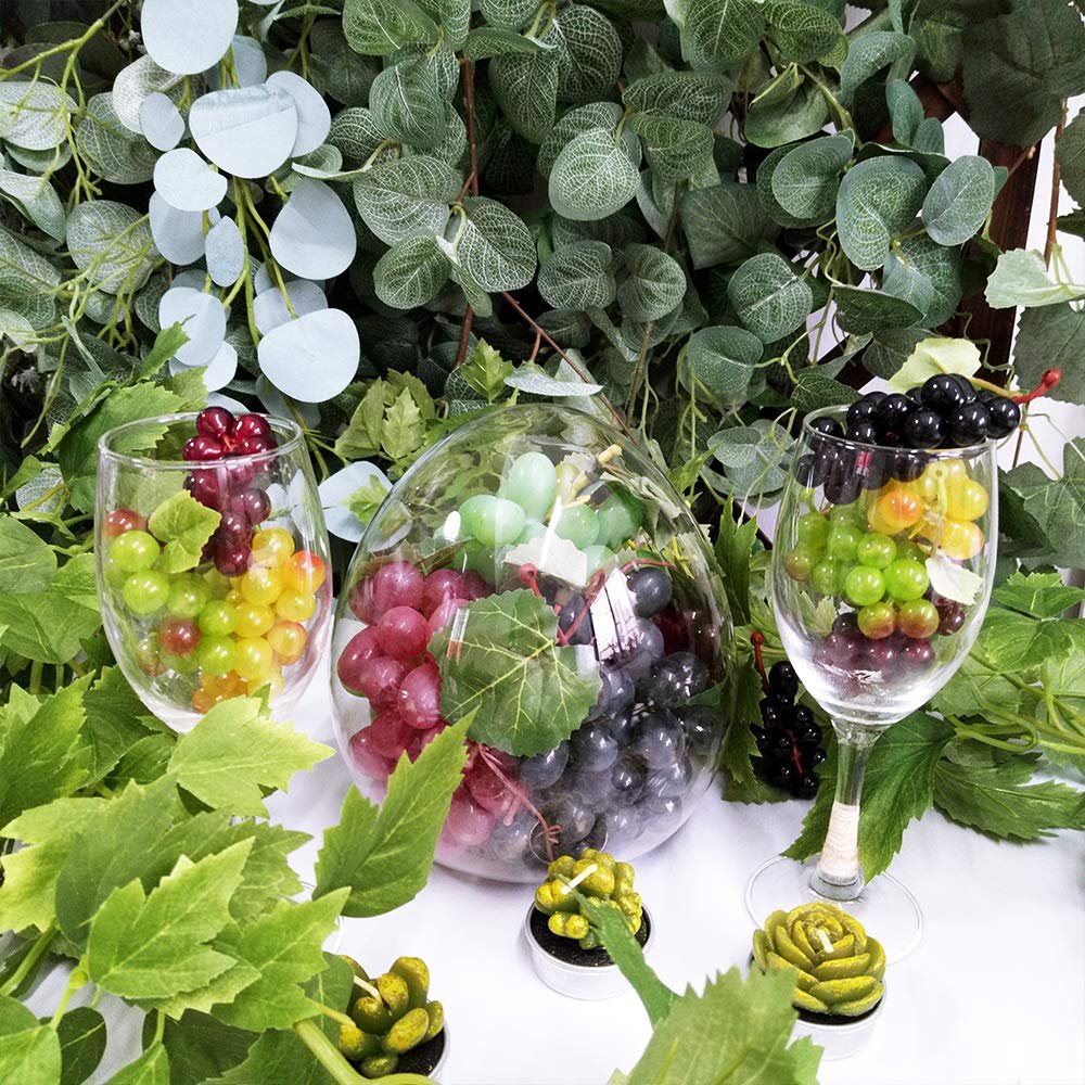 Assorted frosted artificial grape clusters in five colors displayed in glass containers and surrounded by faux greenery.