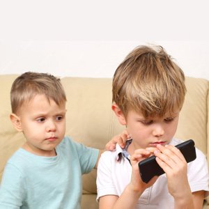 Two young boys sitting on a couch, one holding a smartphone and the other placing a hand on his shoulder.