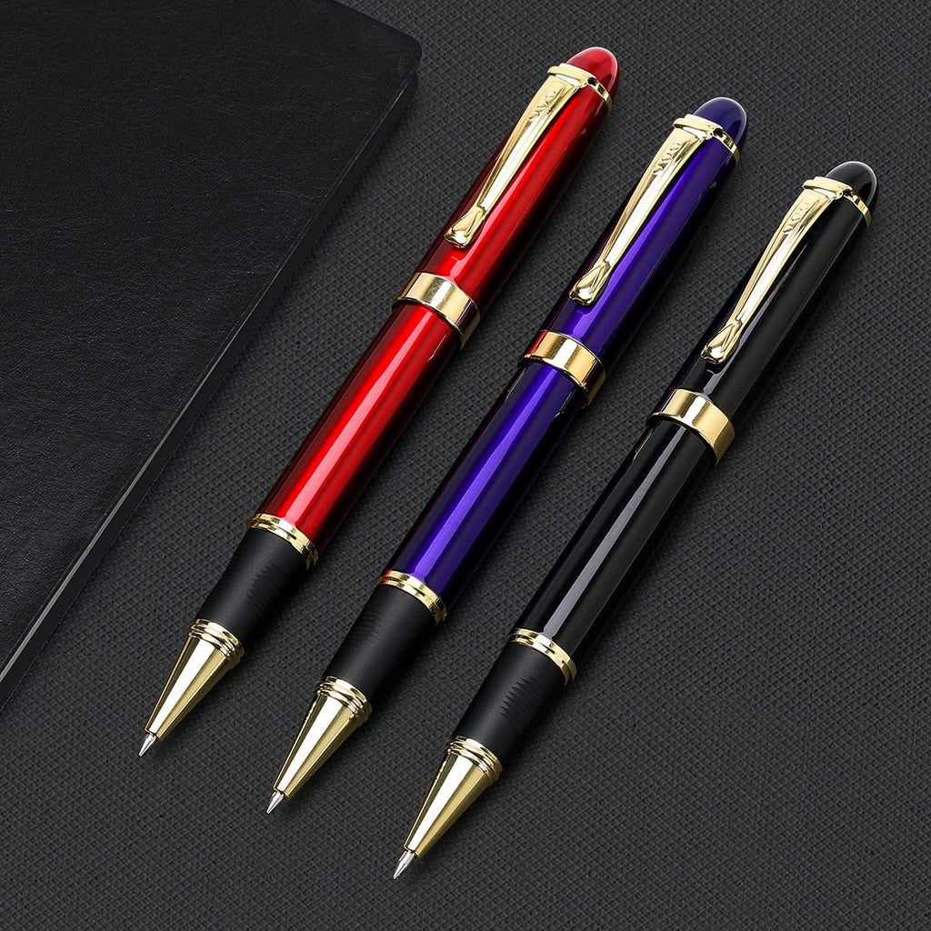 Three metal ballpoint pens in red, purple, and black with gold accents, laid on a dark textured surface next to a black notebook.