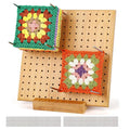 Bamboo crochet blocking board with two colorful granny squares attached by stainless steel pegs, shown at an angle with a wooden base and extra pegs below.