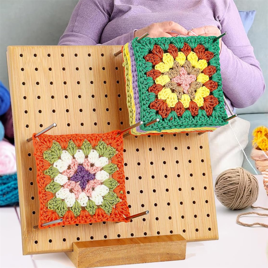 Bamboo crochet blocking board with two colorful granny squares pinned, a person crochets in the background.