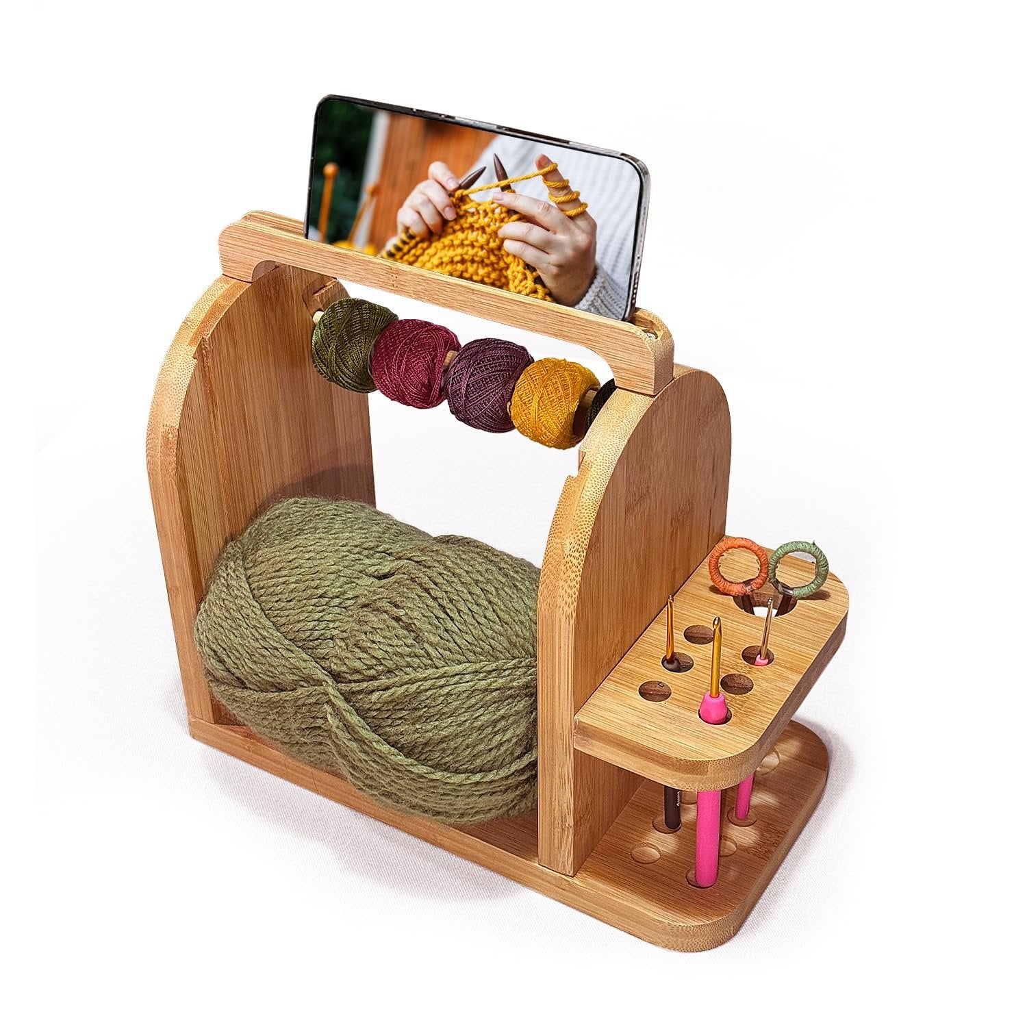 Bamboo yarn organizer with large green yarn ball, four smaller yarn balls on top, and crochet hooks and needles in side slots. Phone stand displays knitting tutorial.