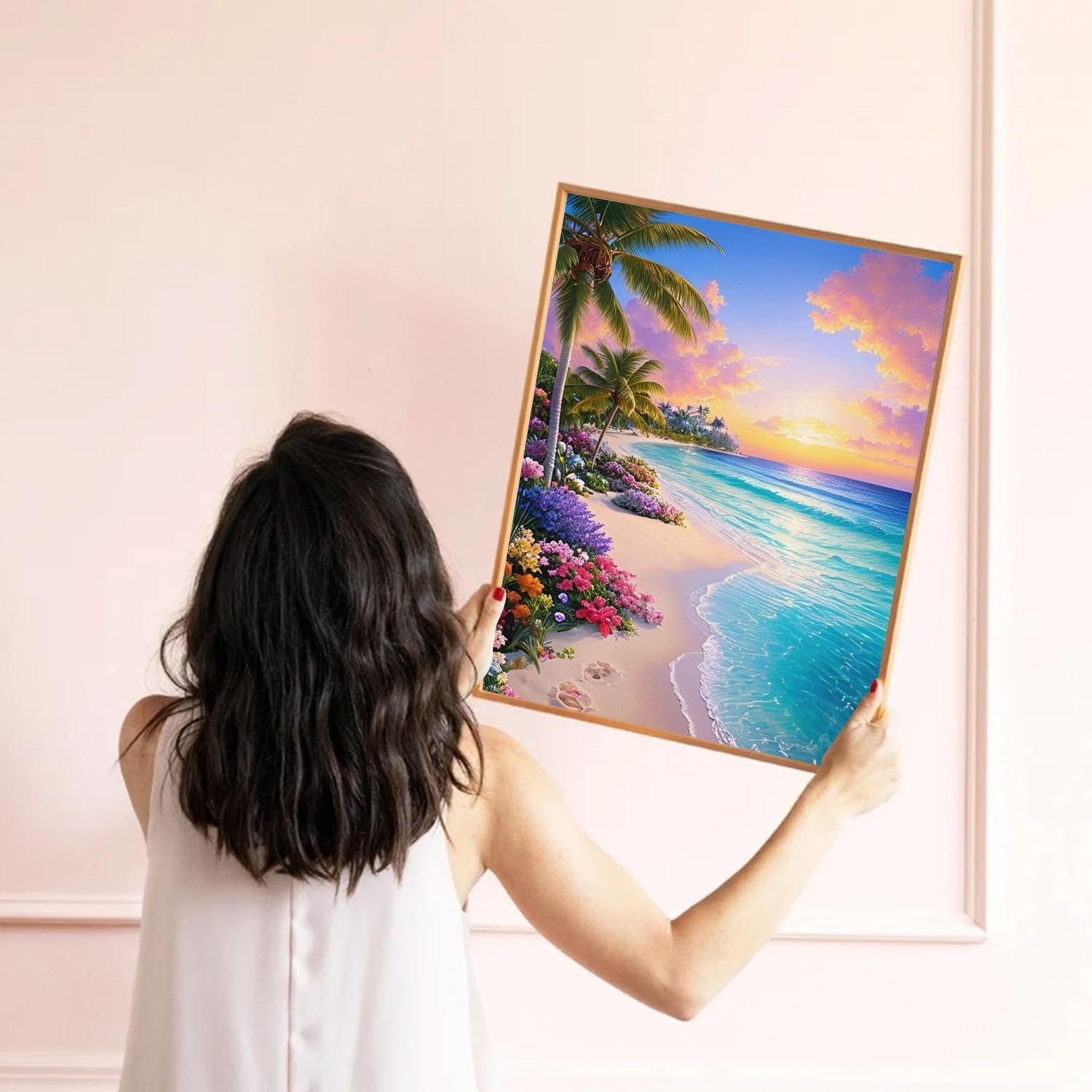 Woman holding a framed diamond art painting of a beach sunset with palm trees and flowers.