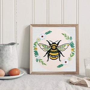 Embroidery hoop art featuring a bumblebee surrounded by flowers and leaves, displayed on a table with eggs and a pitcher.