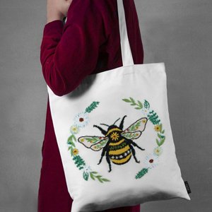 White tote bag with embroidered bee and floral wreath design, held by a person in a maroon shirt.