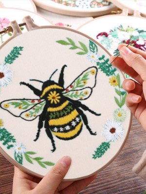 Embroidery hoop with a bee and floral design being stitched by hands, multiple hoops in background