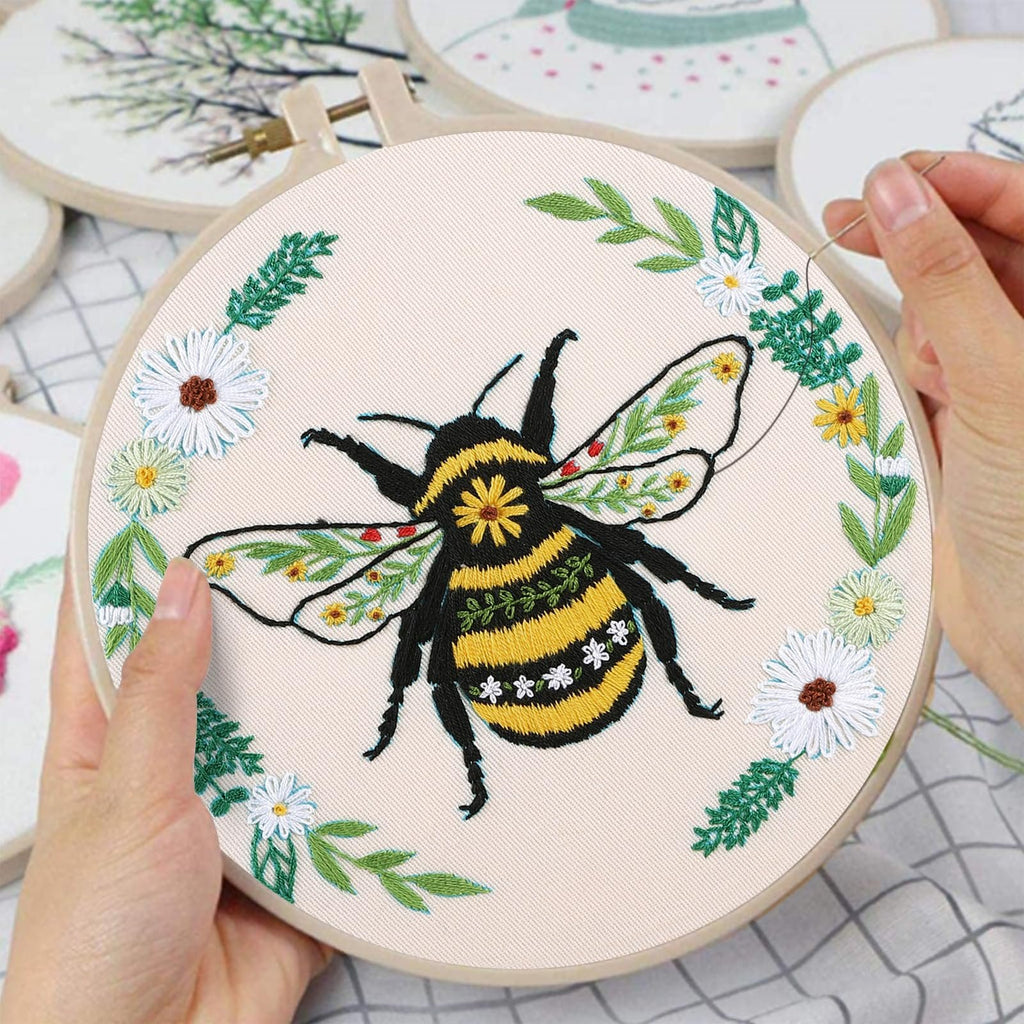 Embroidery hoop with a bumblebee design being stitched by hands, surrounded by floral elements.